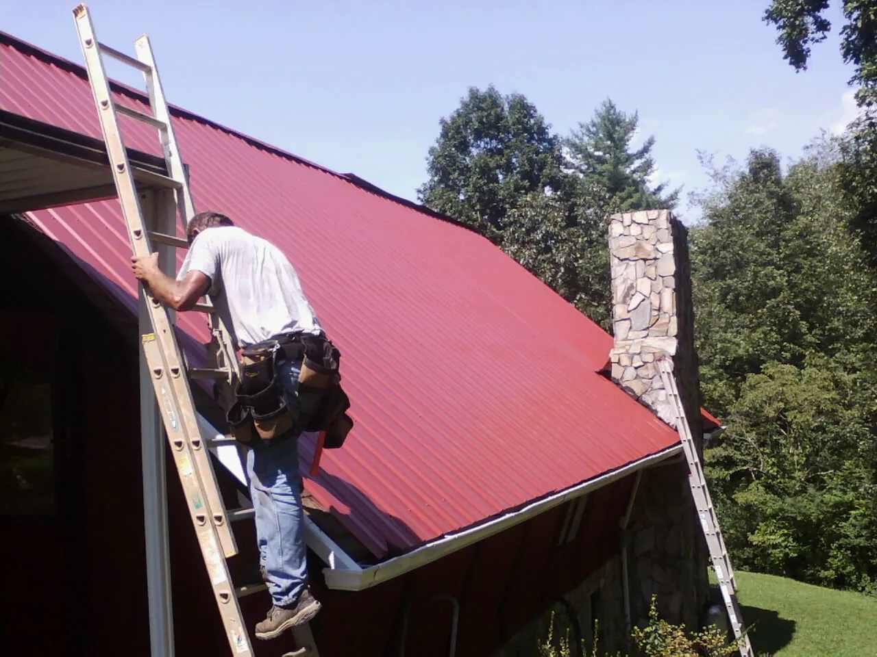 Roofer on a ladder beside a red metal roof and stone chimney — Soffit & Fascia Repair by Roofing Brothers in Barnesville